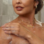 A woman with nude lipstick and gold jewelry gently washes her shoulder with NOPALERA Cactus Shower Gel. Her manicured nails and a “Dominique” necklace are visible against the tiled background.