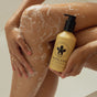 In the shower, a person holding NOPALERA’s Cactus Shower Gel—with a black logo and pump—presses the yellow vegan bottle against their soapy leg, wearing thin gold rings on their fingers.