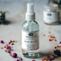 A clear spray bottle labeled Anima Mundi ROSE WATER Beauty Myst, a soothing face toner, rests on marble with dried rose petals and crystals scattered nearby. A second, blurred jar appears in the background.