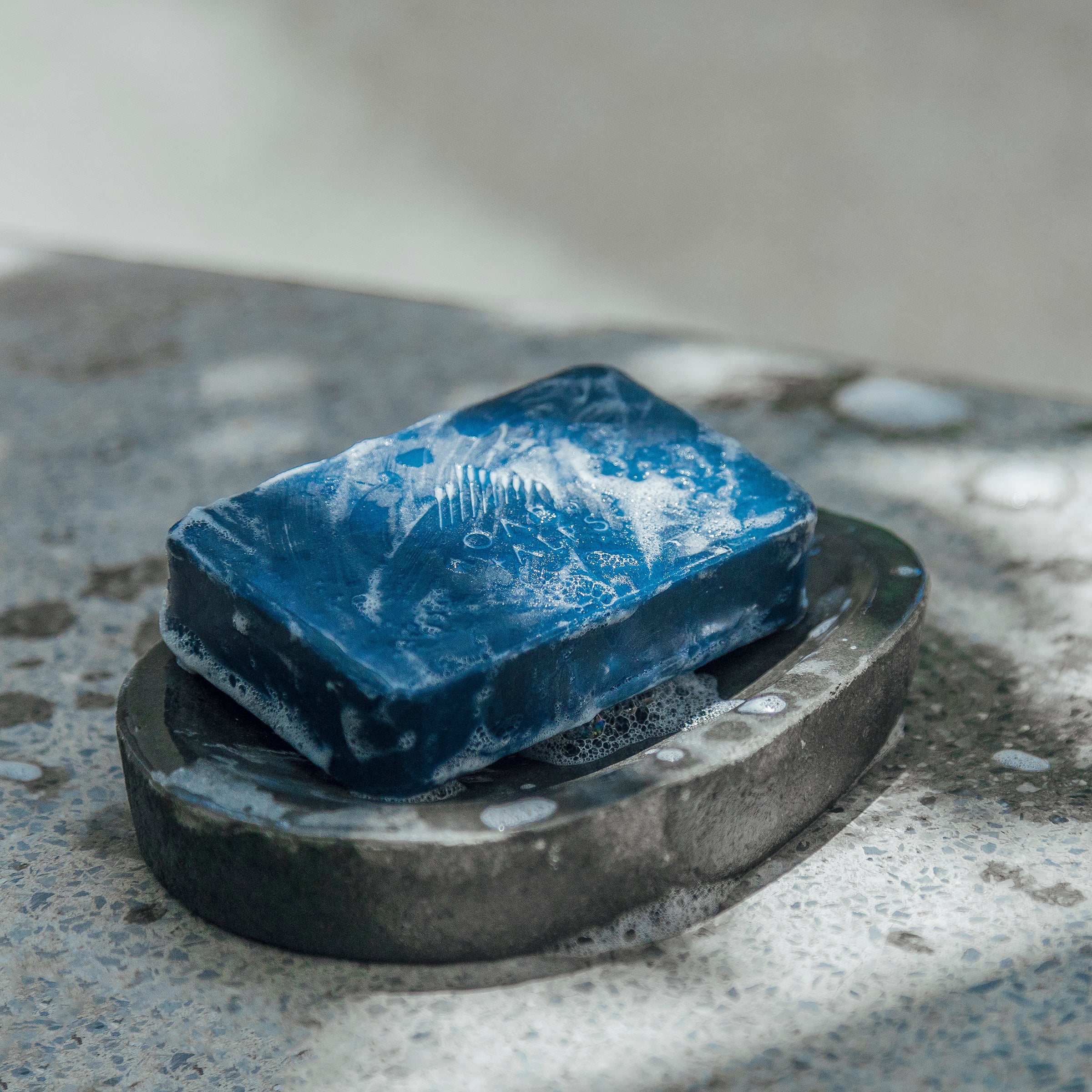 An Exfoliating Bar Soap by Oars + Alps with lather rests on a dark gray soap holder atop a speckled stone surface, where light and shadow patterns enhance its allure.