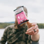 A person in camouflage holds a Kate’s Real Food Organic Energy Bar - Oatmeal Cranberry Almond toward the camera, with a softly blurred lake and trees in the background.