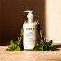 A bottle of Oneka Body Lotion - Rosemary & Mint with a pump rests on a wooden surface, surrounded by fresh rosemary and mint sprigs in warm natural light.