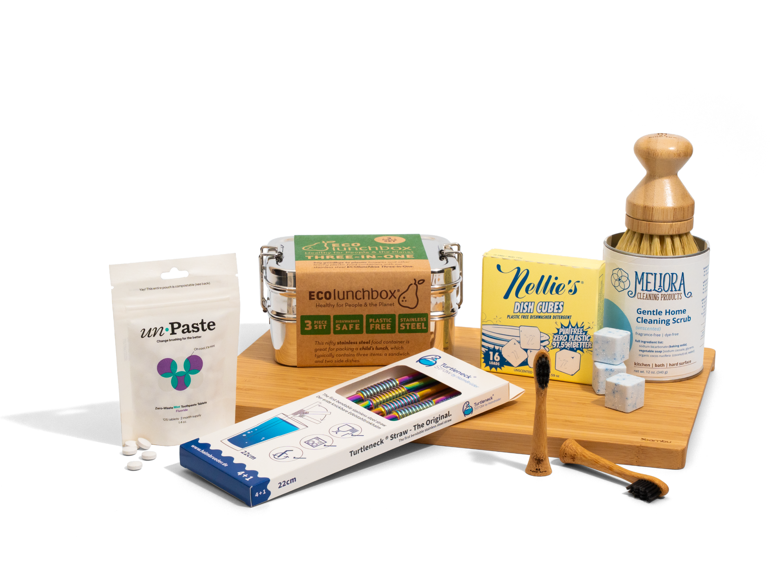 A selection of eco-friendly kitchen products, including a metal lunchbox, toothpaste tablets, dish scrub, cleaning scrub, bamboo toothbrush, colored pencils, and a wooden dish brush, arranged on a light wooden board.