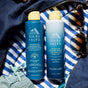 Two Oars + Alps bottles—SPF 50 Hydrating Antioxidant Sunscreen Spray with Niacinamide, and Aloe Cooling Spray—sit on blue and white striped fabric next to sunglasses and a teal rope.