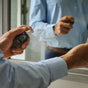 A person in a blue dress shirt sprays Oars + Alps ASCEND Cologne onto their wrist, savoring the refined scent as they stand before a bathroom mirror.