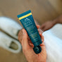 A person holds an open blue tube of Oars + Alps Aluminum-Free Whole Body Cream Deodorant. In the blurred background, there’s a white shoe and a hand on a knee. The formula uses natural starches to absorb moisture effectively.