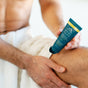 A shirtless man with a towel around his waist applies a blue tube of Oars + Alps Aluminum-Free Whole Body Cream Deodorant to his leg, highlighting his hands, the tube, and his knee. The deodorant is designed for moisture absorption and gentle care.