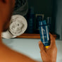 A person holds a blue Oars + Alps Spray Deodorant in front of a bathroom shelf with rolled towels and toiletries in the background.