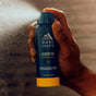 A hand sprays a blue and yellow can of Oars + Alps Spray Deodorant against a blurred brown background.
