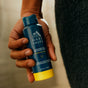 A hand holds an Oars + Alps Spray Deodorant in California Coast scent against a blurred background, possibly wearing a dark towel. This aluminum-free, travel-size deodorant is easy to carry and use on the go.