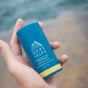A hand holds an Oars + Alps Natural Deodorant stick labeled California Coast with a fresh ocean splash scent, with a blurred outdoor background.