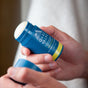 Someone holds an Oars + Alps Natural Deodorant stick in blue, with the cap removed and ready to use. The label shows a mountain logo, white lettering, and advertises a refreshing ocean splash scent.