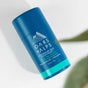 A blue cylindrical stick of Oars + Alps Natural Deodorant in Deep Sea Glacier scent rests on a white surface with a leaf shadow, featuring aluminum-free, odor-preventing formula for long-lasting freshness.
