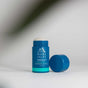A blue stick of Oars + Alps Natural Deodorant, with its cap off, rests on a white surface. The aluminum-free product features a fresh ocean splash scent, while soft shadows and blurred leaves appear in the foreground.
