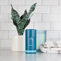 An Oars + Alps Natural Deodorant stick rests on a bathroom counter near a folded towel, potted snake plant, and blue candle, set against white subway tiles—an aluminum-free, odor-preventing pick and Men’s Health Editors Choice.