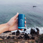 A hand holds an Oars + Alps Natural Deodorant stick in blue packaging in front of a blurred coastal background with rocks, sand, and ocean waves.