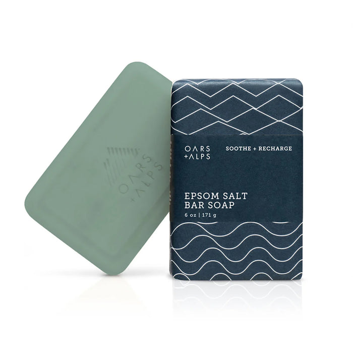 A green Oars + Alps Athlete Bar Soap with Epsom Salt sits beside its dark blue box, decorated with abstract white waves and Soothe + Recharge text, featuring a soothing eucalyptus spearmint scent.