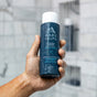 A hand holds a bottle of Oars + Alps Hydrating Shampoo in the shower, water droplets on the skin. The blurred tiled wall in the background emphasizes the shampoo’s hydrating formula.