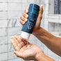 A person squeezes sulfate-free, white Hydrating Shampoo from a dark blue Oars + Alps bottle into their hand in a tiled shower.