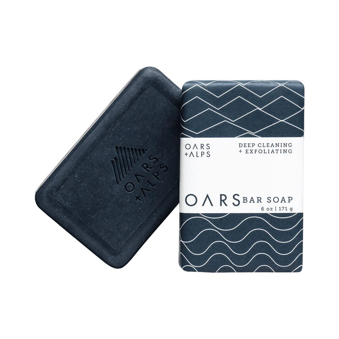 Two Oars + Alps Exfoliating Bar Soaps: one unwrapped, dark blue with an embossed logo; the other wrapped in blue paper with white geometric patterns and labeled Deep Cleaning + Exfoliating with volcanic sand.