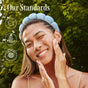 A smiling woman with a blue headband uses Three Ships Off Duty Sea Algae + Bio-Peptide Smoothing Eye Masks to cleanse her face outdoors. Three certification icons and Our Standards appear above, with green trees in the background.