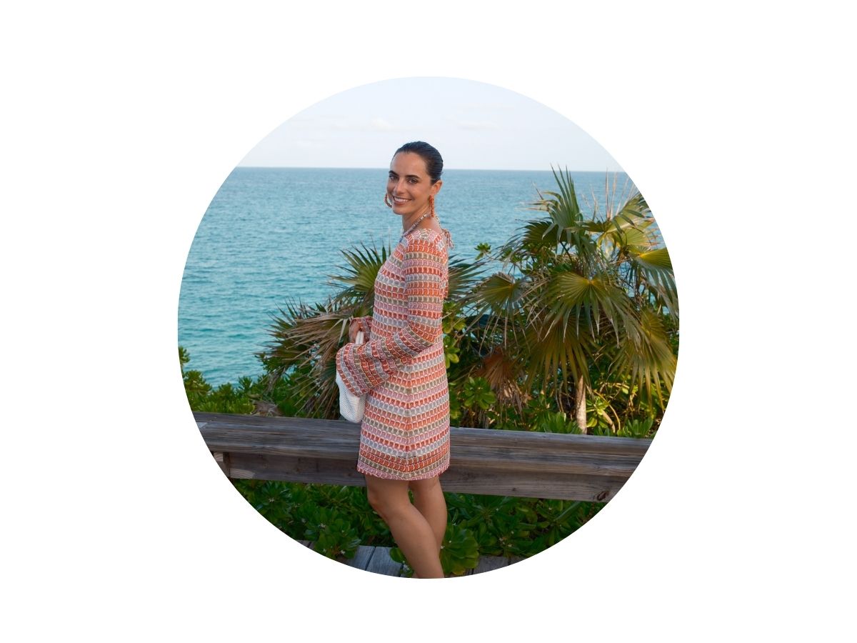 A woman in a patterned dress stands by a wooden railing, smiling at the camera with the ocean and palm trees in the background on a sunny day.