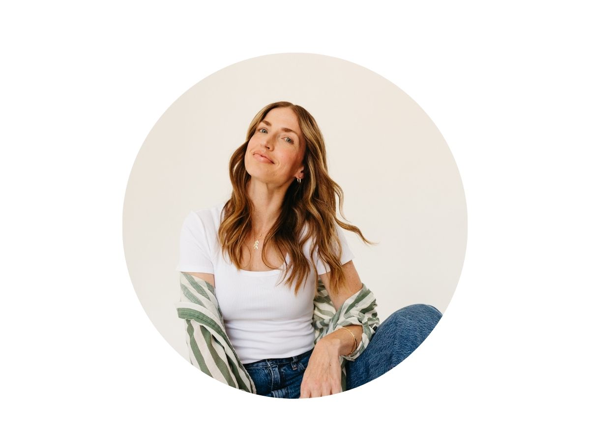A woman with long, wavy brown hair is sitting and smiling slightly. She is wearing a white t-shirt, jeans, and a green striped shirt draped over her arms, with a plain light background.