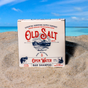 A box of Old Salt Hair/Beard Shampoo rests on a sandy beach against turquoise sea and blue sky. The cream-colored packaging features red and blue text plus nautical graphics.
