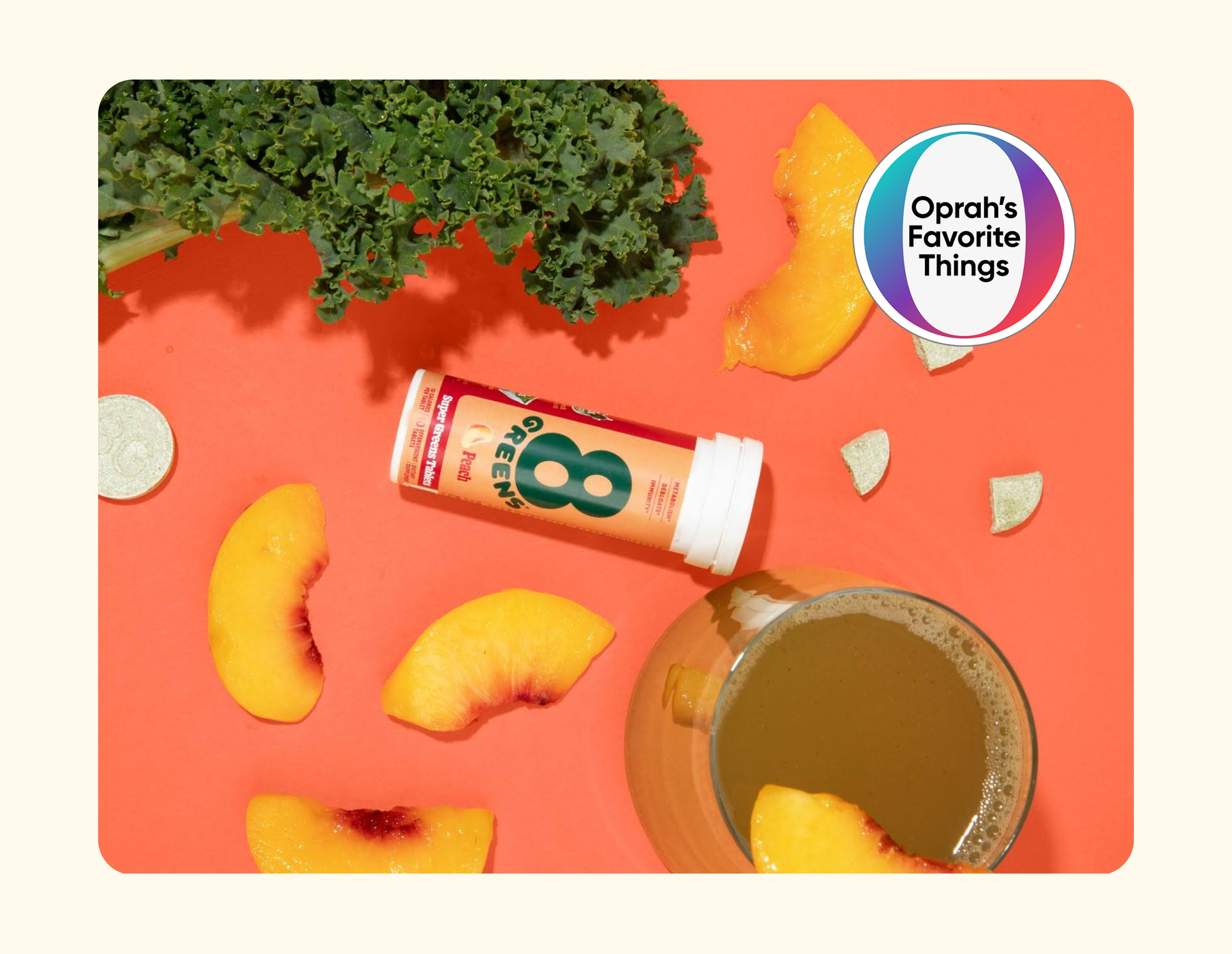 A tube of 8Greens tablets lies on a red surface surrounded by kale, peach slices, and a glass of green juice. A coin and small citrus wedges are nearby. An Oprahs Favorite Things badge is in the top right corner.