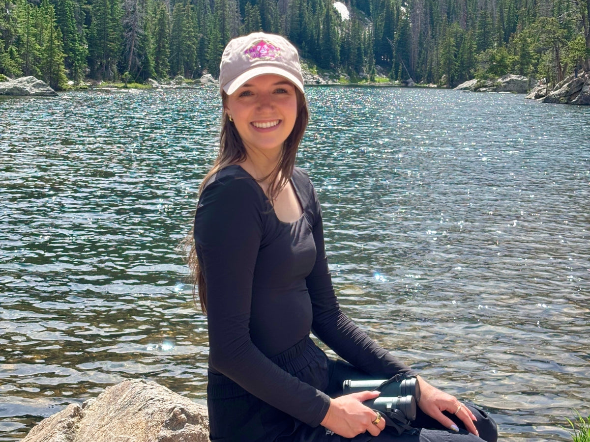 A young woman in a black outfit and light-colored cap sits on a rock by a lake, smiling, with trees in the background. She holds binoculars and sunlight reflects on the water behind her.