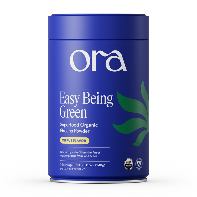 A blue cylindrical Ora Organic container labeled "Easy Being Green: Daily Active Superfood Greens Powder, Citrus Flavor," features green leaf graphics, certification icons, and highlights organic prebiotics on the front.