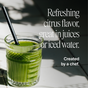 A glass of green juice with a black straw sits on a sunlit white surface. Text beside it reads: Easy Being Green by Ora Organic—delicious citrus taste, boosted with superfood greens. Perfect for juices or iced water. Chef-crafted.