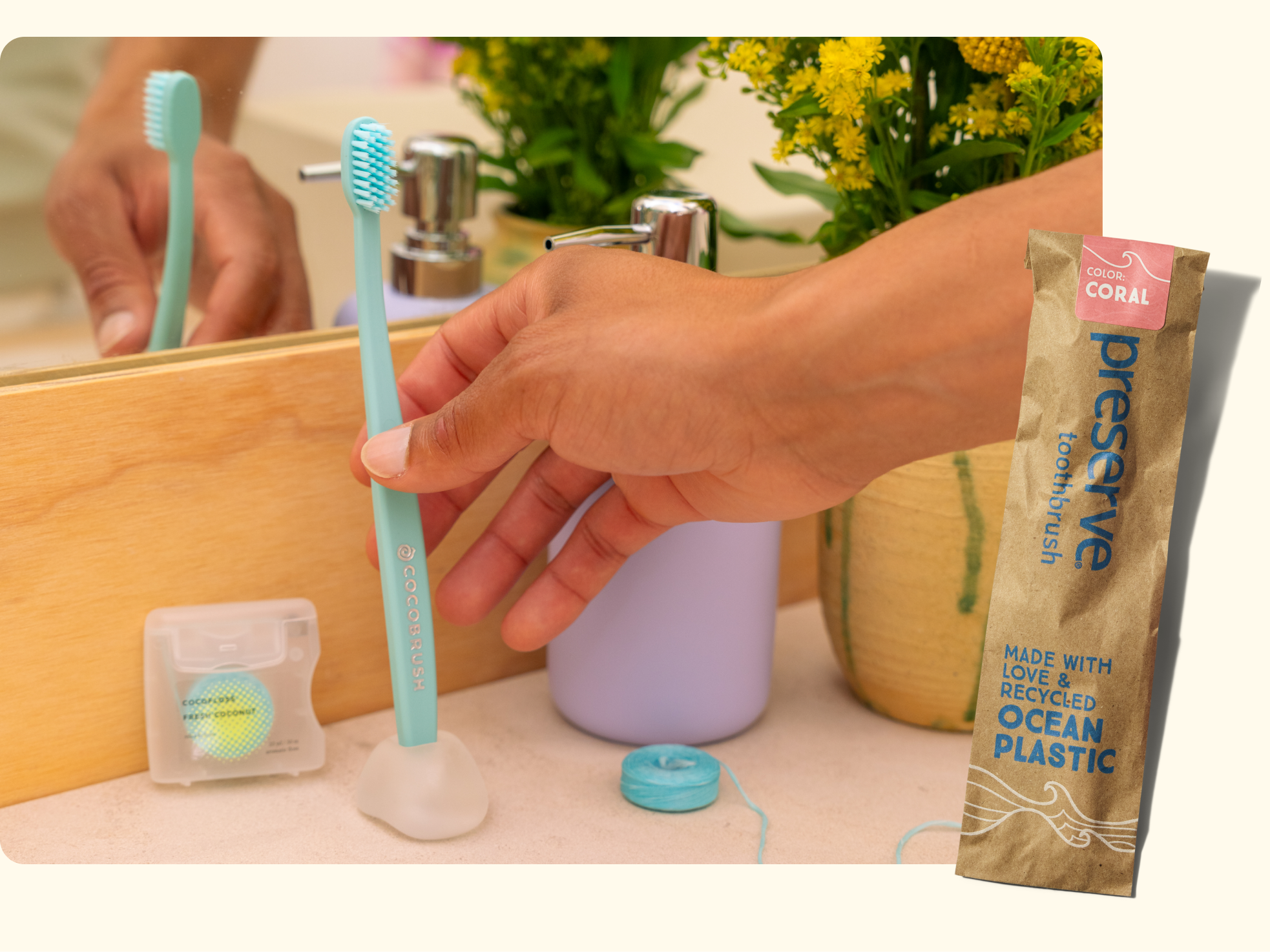 A hand holds a blue toothbrush next to dental floss and a purple soap dispenser on a bathroom counter, with yellow flowers and a Preserve toothbrush package labeled made with recycled ocean plastic.