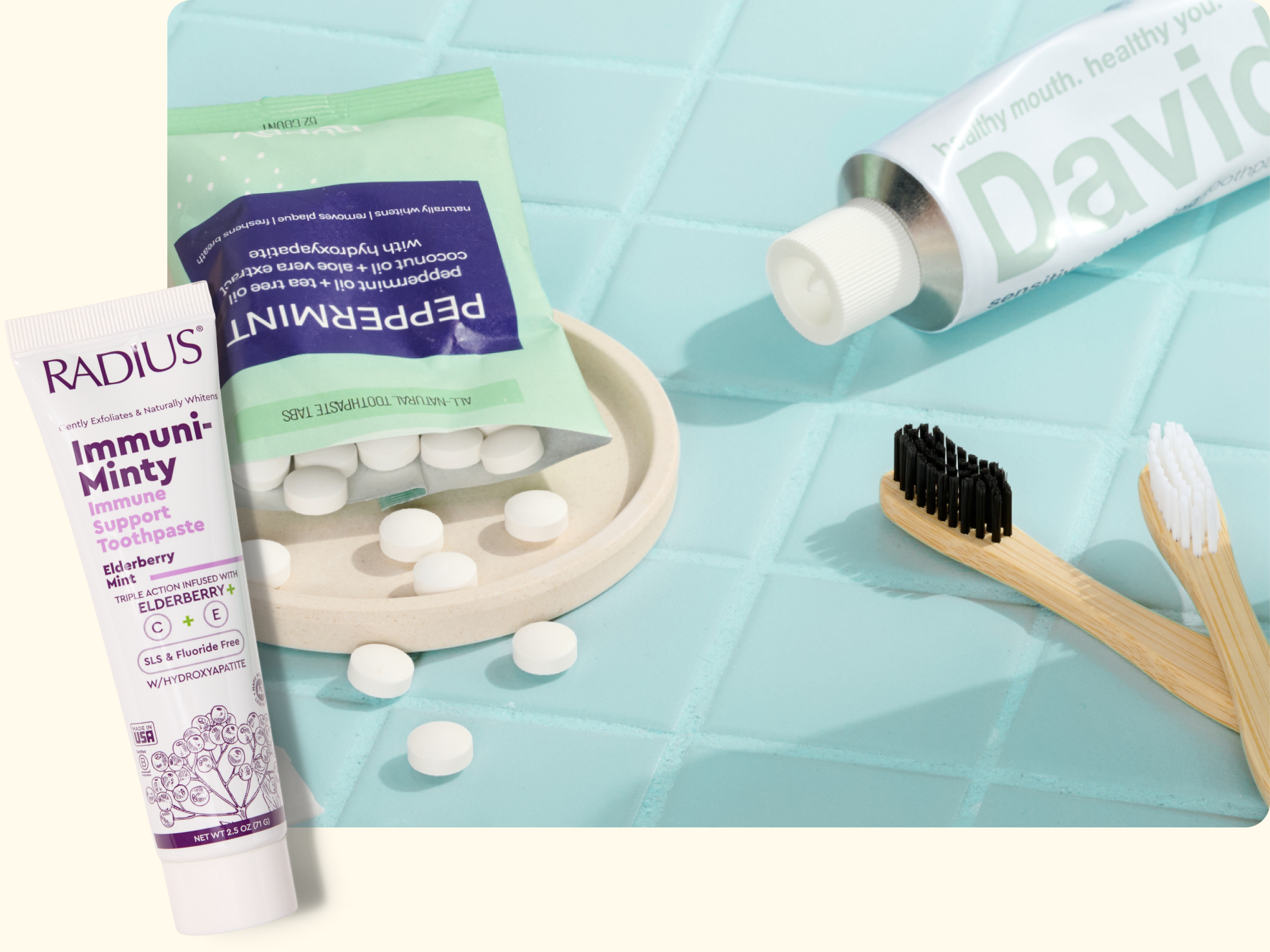 A tube of RADIUS Immuni-Minty toothpaste, a pack of PEPPERMINT mints with tablets spilled out, a tube of DAVIDS toothpaste, and two bamboo toothbrushes are arranged on a blue tile and cream background.