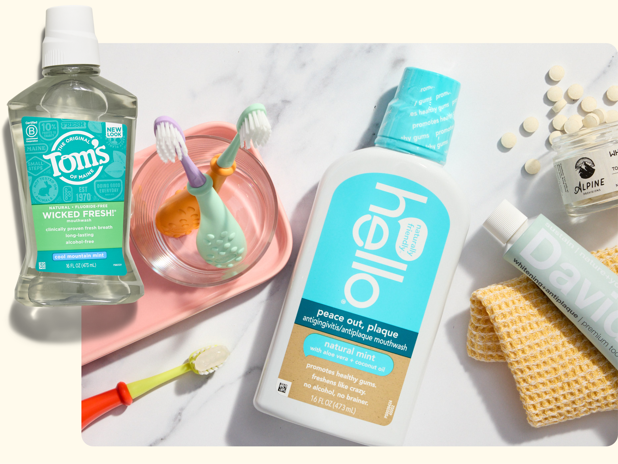 A flat lay of oral care products including Tom’s mouthwash, Hello mouthwash, a Davids toothpaste tube, a baby toothbrush holder with two toothbrushes, a yellow cloth, and a small jar of chewable tablets.