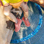 A toddler in a yellow shirt and orange shorts sits by a pool, their legs in the water. The bright pink water shoes match their Green Sprouts Eco Snap Swim Diaper made from recycled polyester. Blue mosaic tiles line the pools edge as sunlight casts playful shadows on the scene.