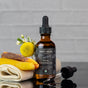 A bottle of Chagrin Valley Soap & Salve Facial Cleansing Oil Makeup Remover with a dropper rests on a folded cloth near yellow flowers and beeswax bars, set against a light gray brick background—ideal for all skin types.
