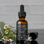 A brown glass dropper bottle of Chagrin Valley Soap & Salve Facial Oil stands upright with its dropper cap beside it. Small white flowers and greenery are arranged nearby on a gray surface.