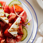 A close-up of a Greek salad with tomatoes, cucumbers, red onions, and feta cheese triangles drizzled with Kosterina Original Balsamic Vinegar on a white plate with a blue rim.