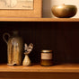 A wooden shelf with a small brown ceramic jug, a tiny vase holding two feathers, a brown soy candle, and a round, lidded brass container. Part of a framed artwork is visible above the shelf.