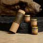 A cylindrical brown box from P.F. Candle Co. rests beside three amber glass jars with gold lids, each labeled as the Greatest Hits Mini Candle Gift Set, 3.5 oz, set against a rustic background for a cozy, natural vibe.