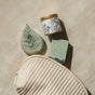 Flat lay of Pacha Soap Co.’s Self Care Spa Set - Vanilla & Sage, showcasing a quilted beige pouch, green leaf bath bomb, blue and cream floral candle, and rectangular green soap on a light textured surface.