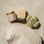 A Self Care Spa Set - Santal & Amber by Pacha Soap Co. featuring a ribbed cream pouch, a floral tin, a woodsy Santal & Amber bar soap, and a glittery cactus bath bomb is displayed on a light beige surface.