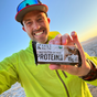 A smiling man in a neon green jacket, cap, and sunglasses holds up a Kate’s Real Food Organic Protein Bar - Peanut Butter Brownie outdoors under a bright sky.