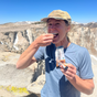 A person in a blue shirt and tan cap smiles while enjoying a Kates Real Food Organic Protein Bar - Peanut Butter Cup on a rocky mountain summit under a clear blue sky, with snow patches and distant peaks in the background.