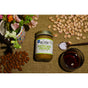 A jar of Big Spoon Roasters Pistachio Crunch Almond Butter, featuring California pistachios, sits among almonds, salt, a glass of dark liquid, and a floral mug on a woven brown surface.