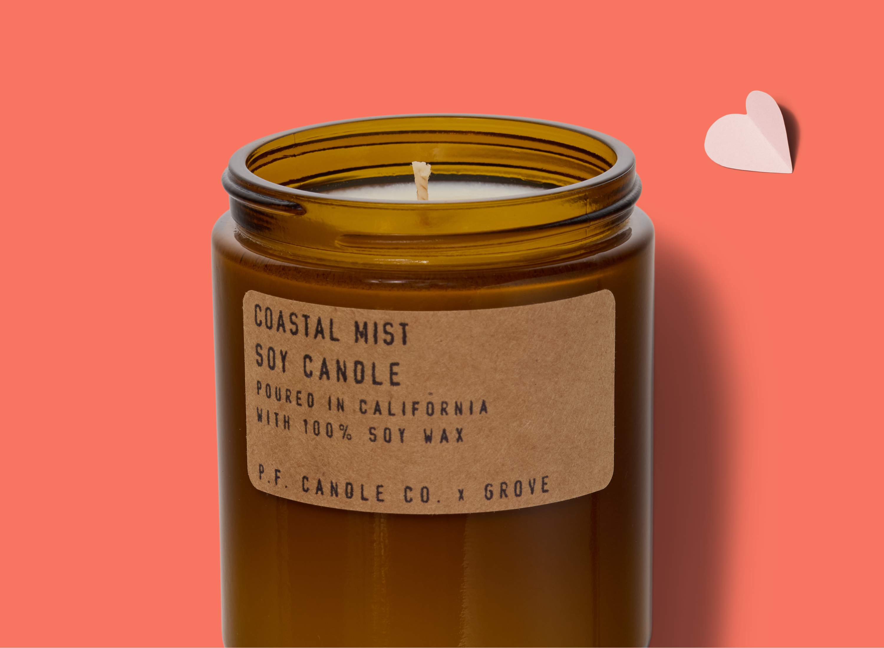 A brown glass jar candle labeled Coastal Mist Soy Candle sits on a coral background with a small paper heart beside it. The label notes it is made in California with 100% soy wax by P.F. Candle Co.