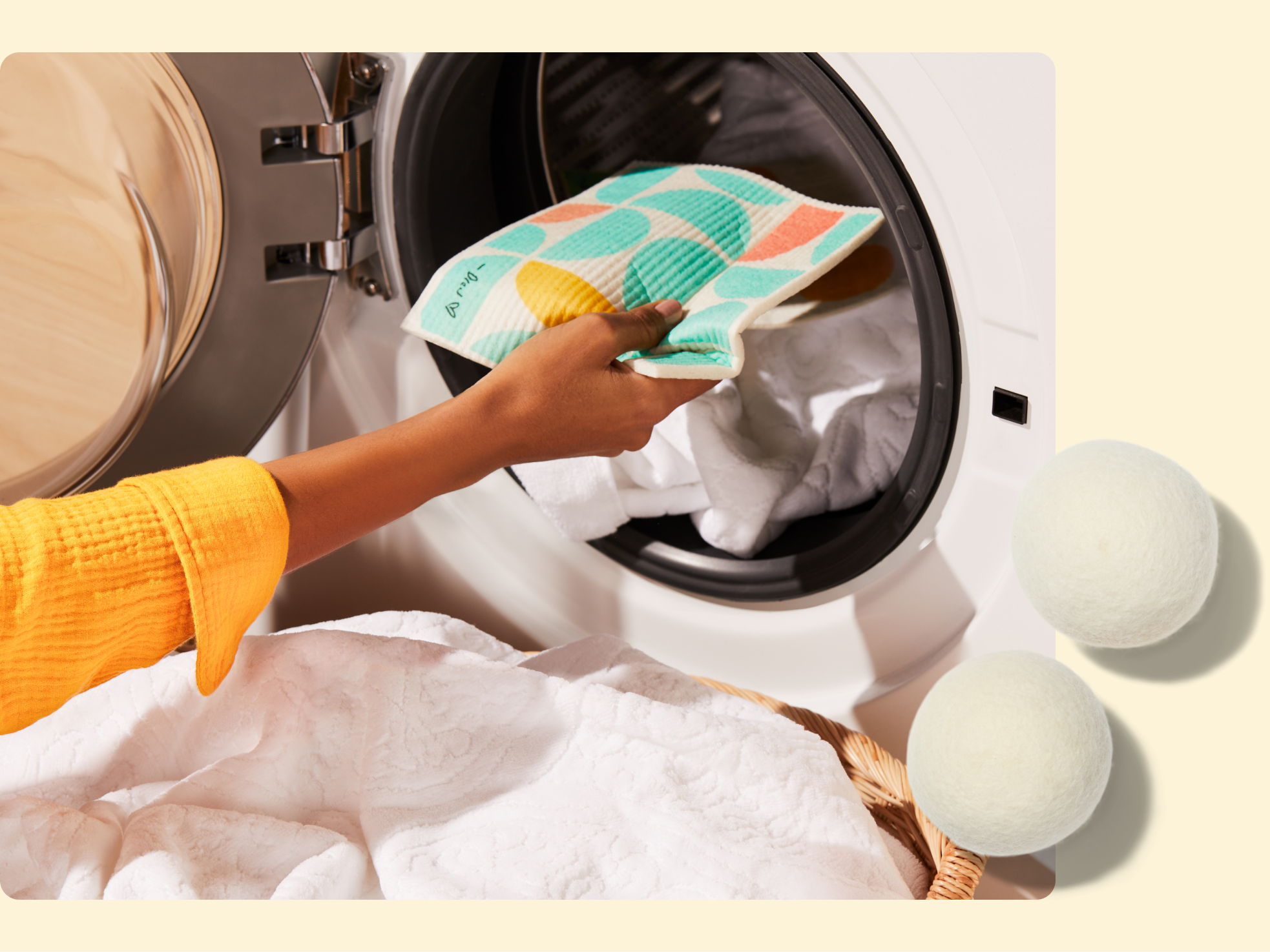 A person in a yellow sleeve places a patterned fabric pouch into a front-loading washing machine filled with white laundry; two white wool dryer balls are shown to the side.