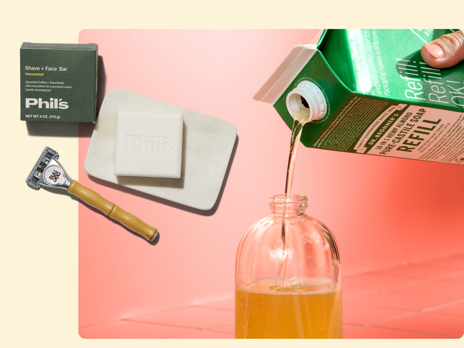A hand pours liquid soap from a green refill carton into a clear bottle. Nearby are a white Phil’s shave and face bar on a dish and a razor with a wooden handle, all on a pink and cream background.