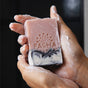 A close-up of soapy hands holding a pink and black marbled Pacha Soap Co. Bar Soap with the brand’s name and a sunburst design engraved on it.